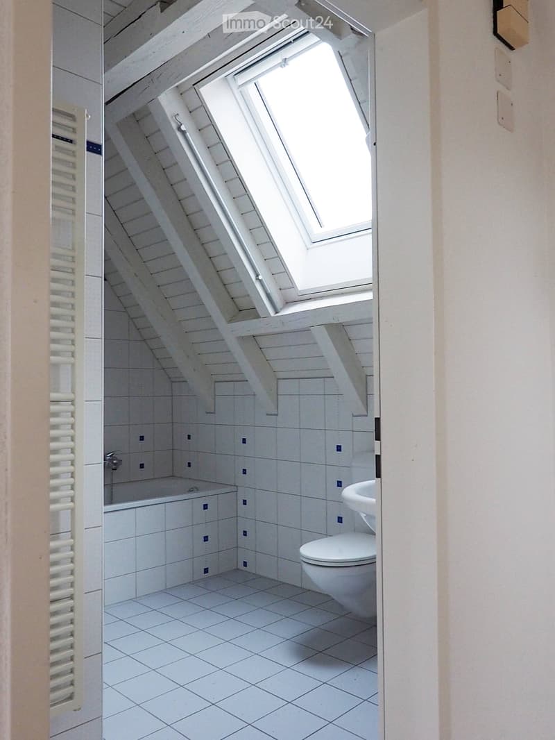 Bright, compact bathroom with white tiled walls and floor, featuring a toilet, bathtub, and a window with a skylight above.