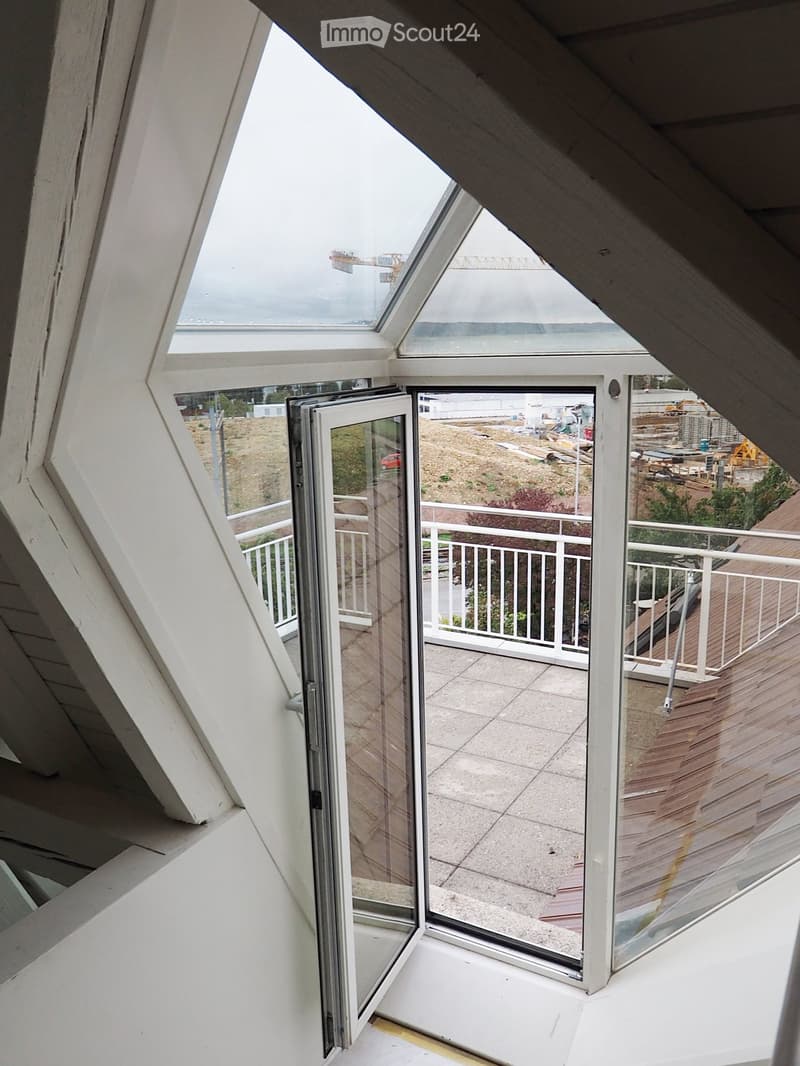 Open sliding glass doors lead to a balcony with a wooden deck and white railing, overlooking a construction site.