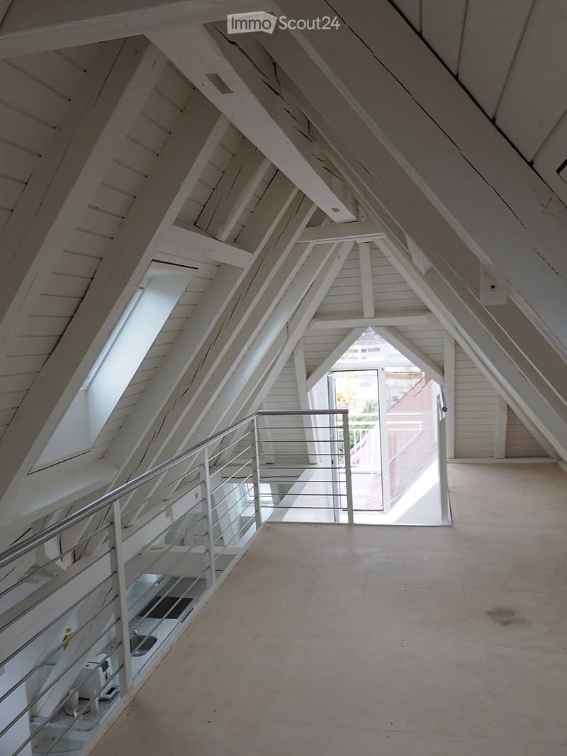 Bright, open attic corridor with exposed wooden beams, white walls, and a modern metal railing leading to a staircase. Natural light streams in from skylights and a large window at the end.