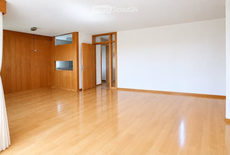 Empty room with light wooden flooring, white walls, and a wooden door leading to another space. The room has a modern, minimalist design with a visible light fixture on the ceiling.