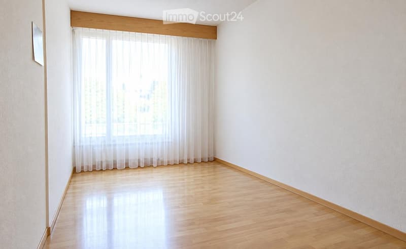 Empty room with wooden flooring, white walls, and sheer curtains covering a large window.