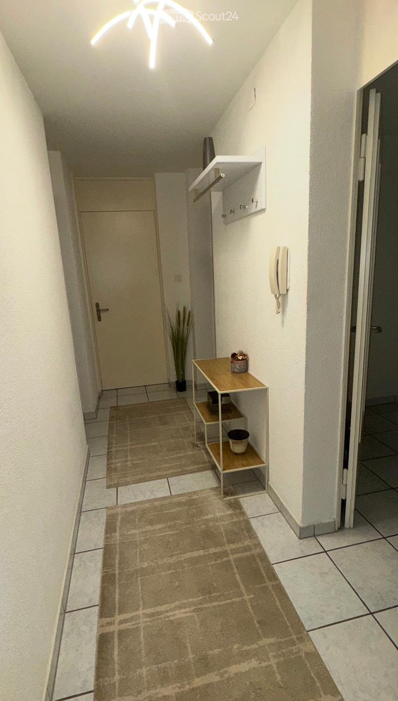 A narrow hallway with tiled flooring, white walls, and a small wooden shelf with decorative items. There is a door on the left and a telephone on the right wall.