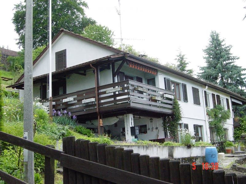 Two-story white house with a balcony, surrounded by greenery and a wooden fence.