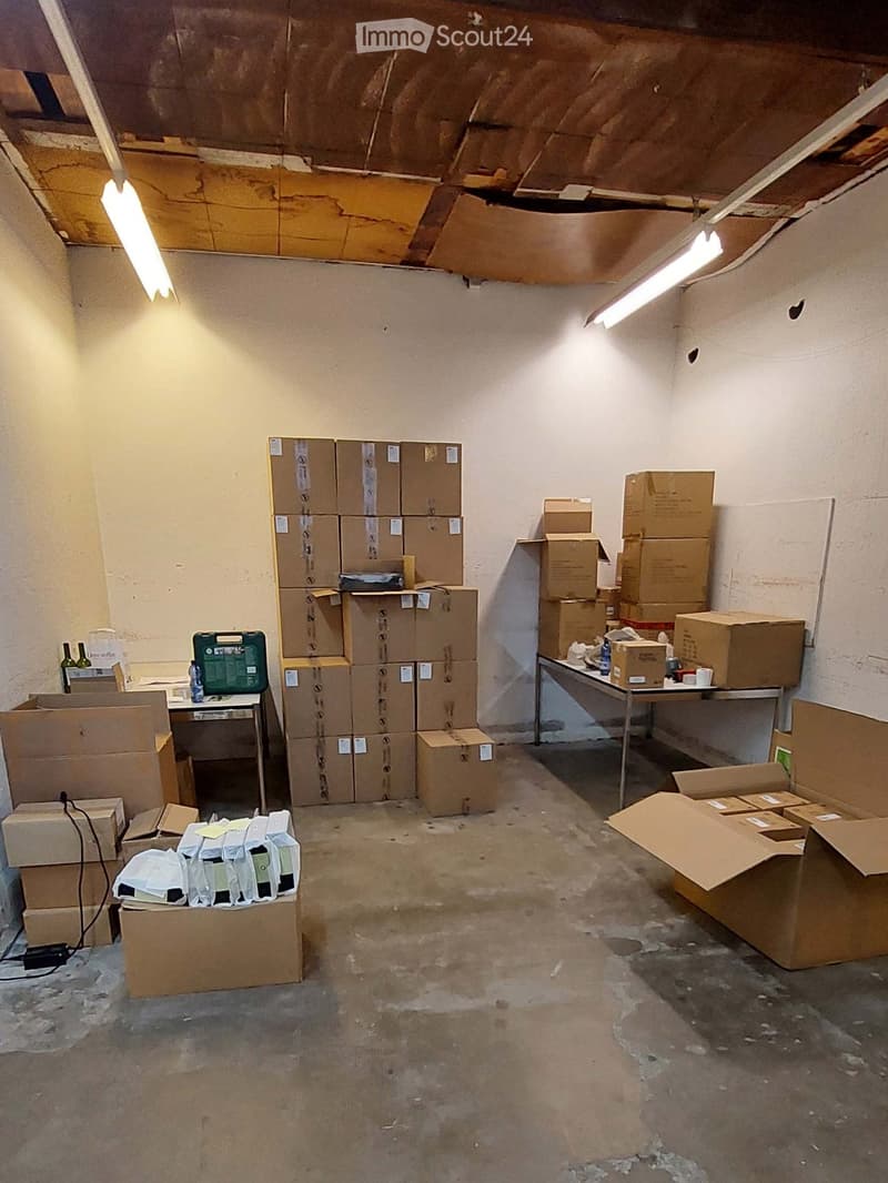 A storage room with exposed ceiling beams, fluorescent lighting, and numerous cardboard boxes stacked throughout the space. A small table with a chair and a few bottles is visible on the left side.