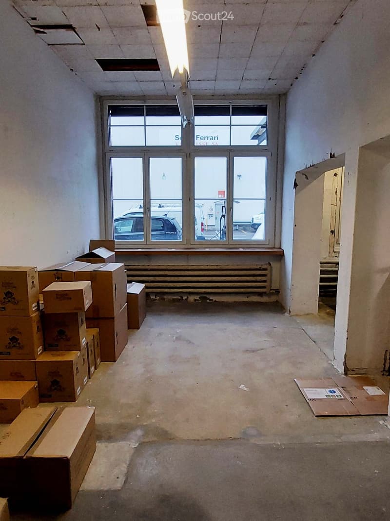 Empty room with bare walls, concrete floor, and large windows showing a parking lot outside. Several cardboard boxes are stacked on the left side.