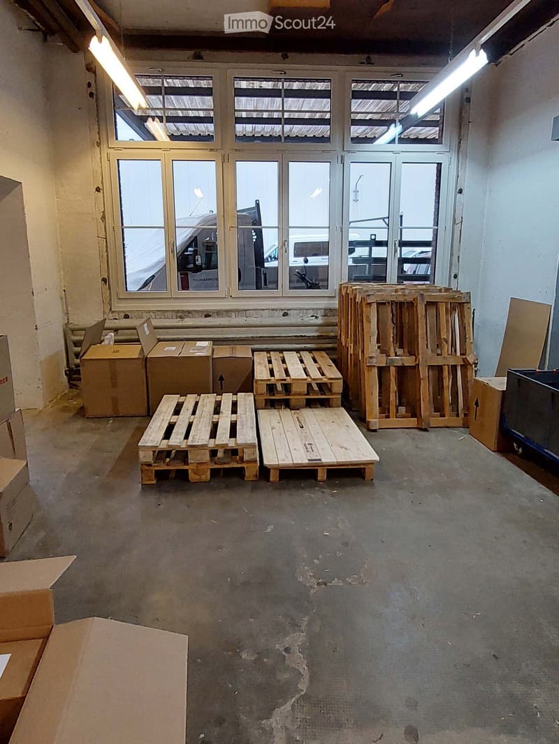 Empty room with concrete floor, large windows, and wooden pallets and cardboard boxes stacked around the perimeter.