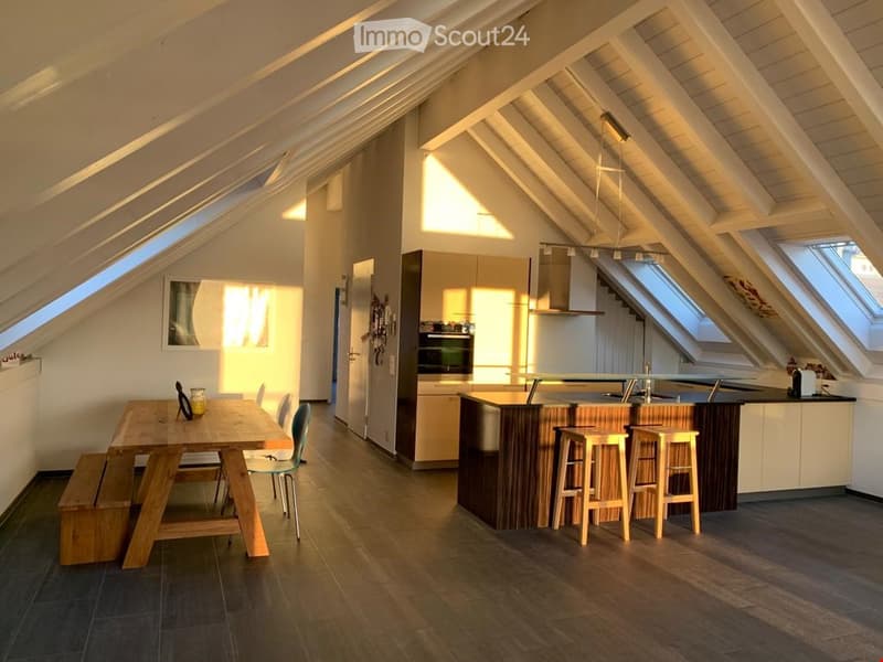 Bright attic space with wooden beams, featuring a kitchen area with an island, stools, and a dining table with chairs and a bench.