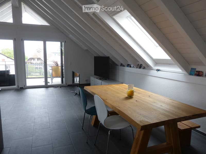 Bright attic room with wooden table, chairs, TV, and large windows, featuring a modern design and ample natural light.