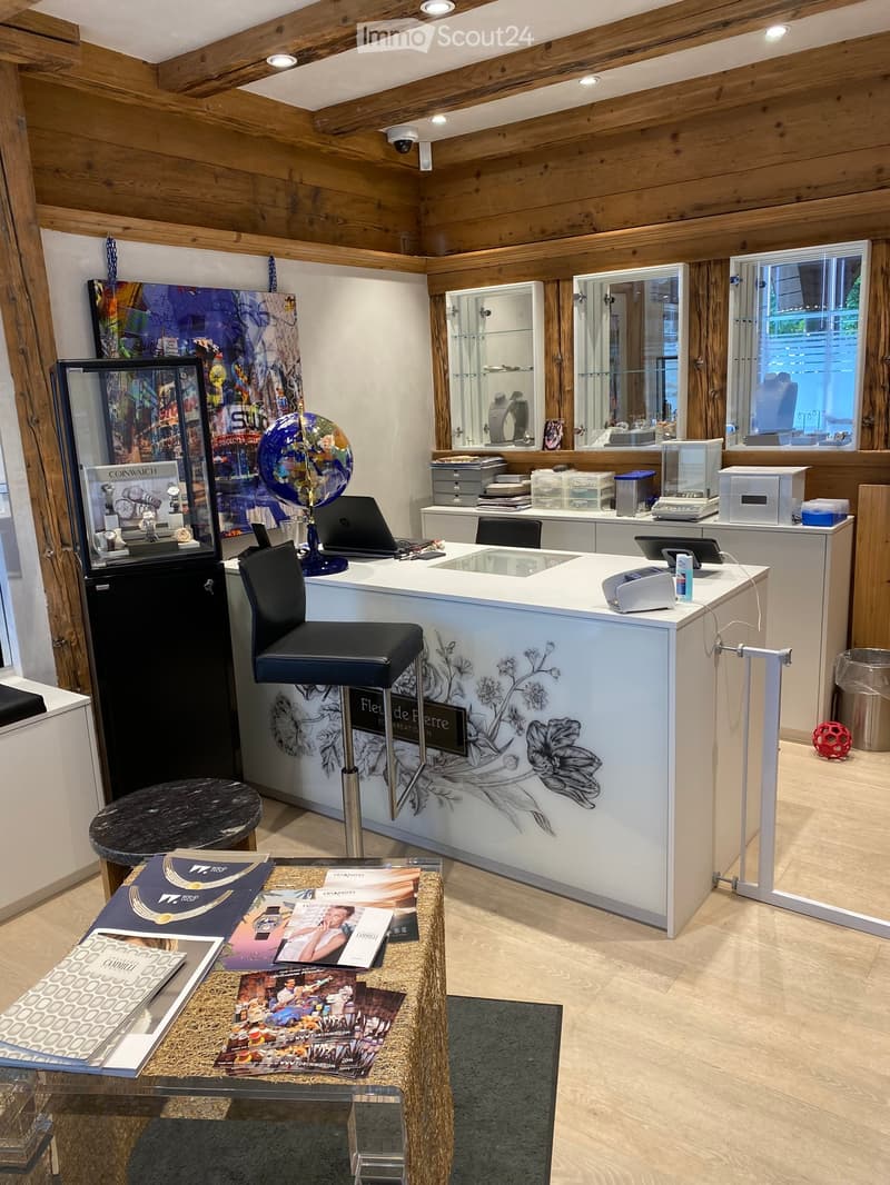 A modern, well-lit interior with wooden beams and white walls, featuring a reception area with a white counter, a black stool, a laptop, a globe, a mirror, and a display case with watches. There are brochures on a small table and a black display case with