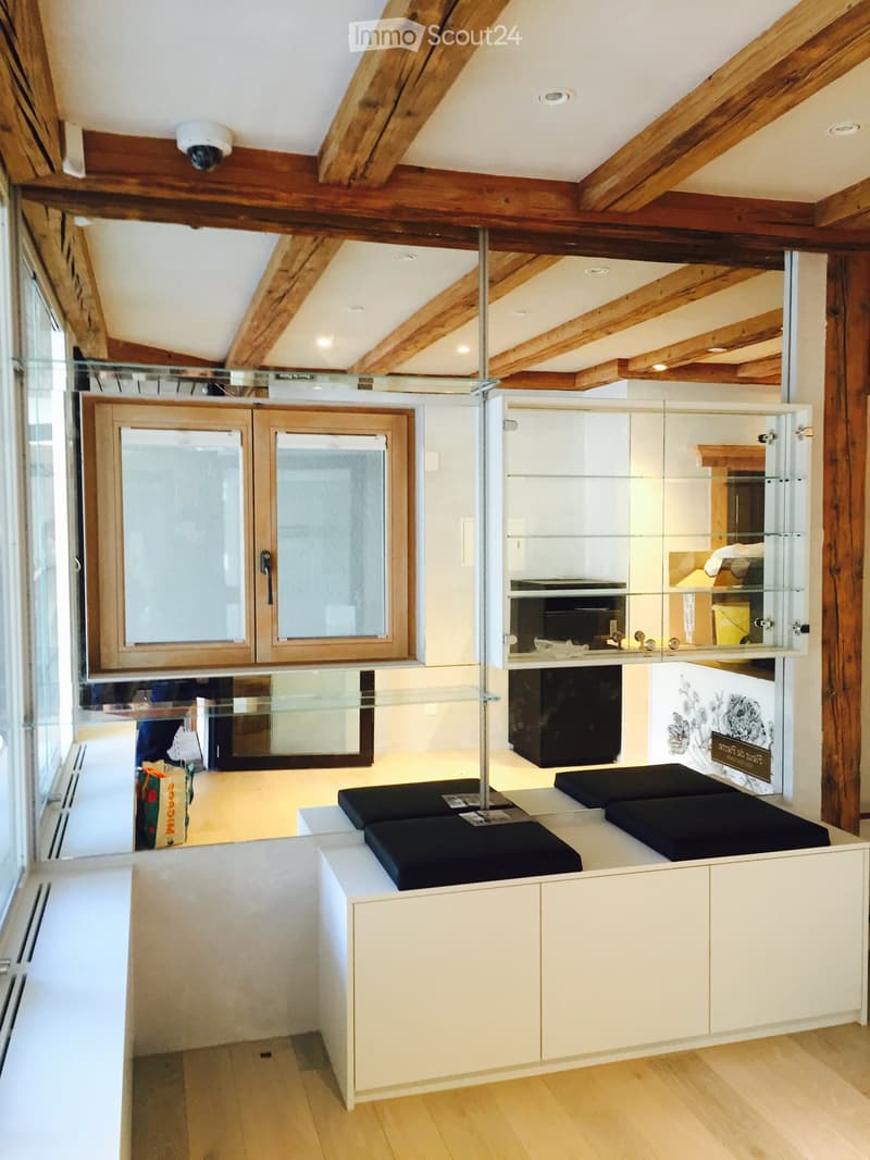 Modern, minimalist room with exposed wooden beams, white cabinets, glass display case, and black stools.