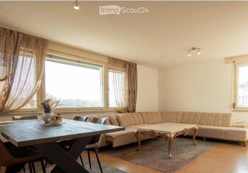 Modern living room with large windows, beige sectional sofa, coffee table, dining table, and decorative vase on the table.