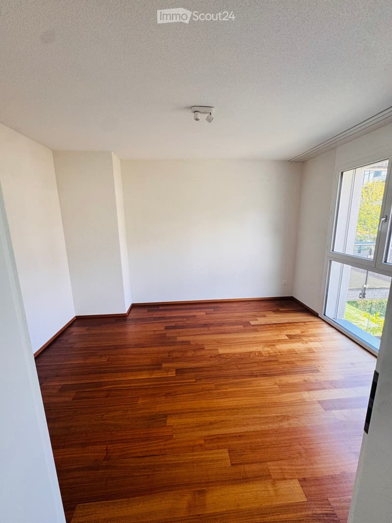 Empty room with wooden flooring, white walls, and a large window with a view of greenery outside.