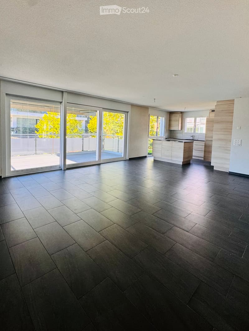 Empty room with dark tiled floor, large sliding glass doors leading to a balcony, and a small kitchen area visible in the background.