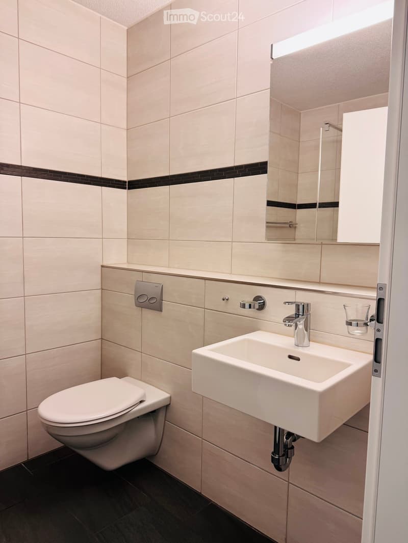 Modern bathroom with white tiles, a toilet, a sink with a chrome faucet, a mirror, and a glass cup on the wall.