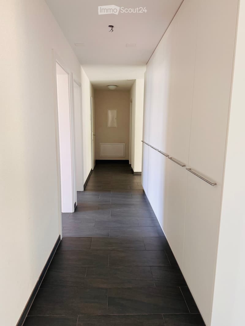 Clean, modern hallway with dark tiled flooring, white walls, and a metal handrail on one side. Features recessed ceiling lighting and several doors leading to adjacent rooms.