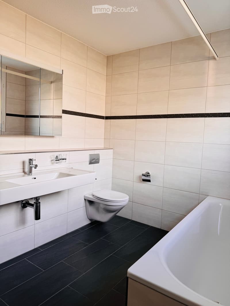 Modern bathroom with white tiles, black floor tiles, a white sink, a toilet, a bathtub, and a mirror with a built-in shelf.