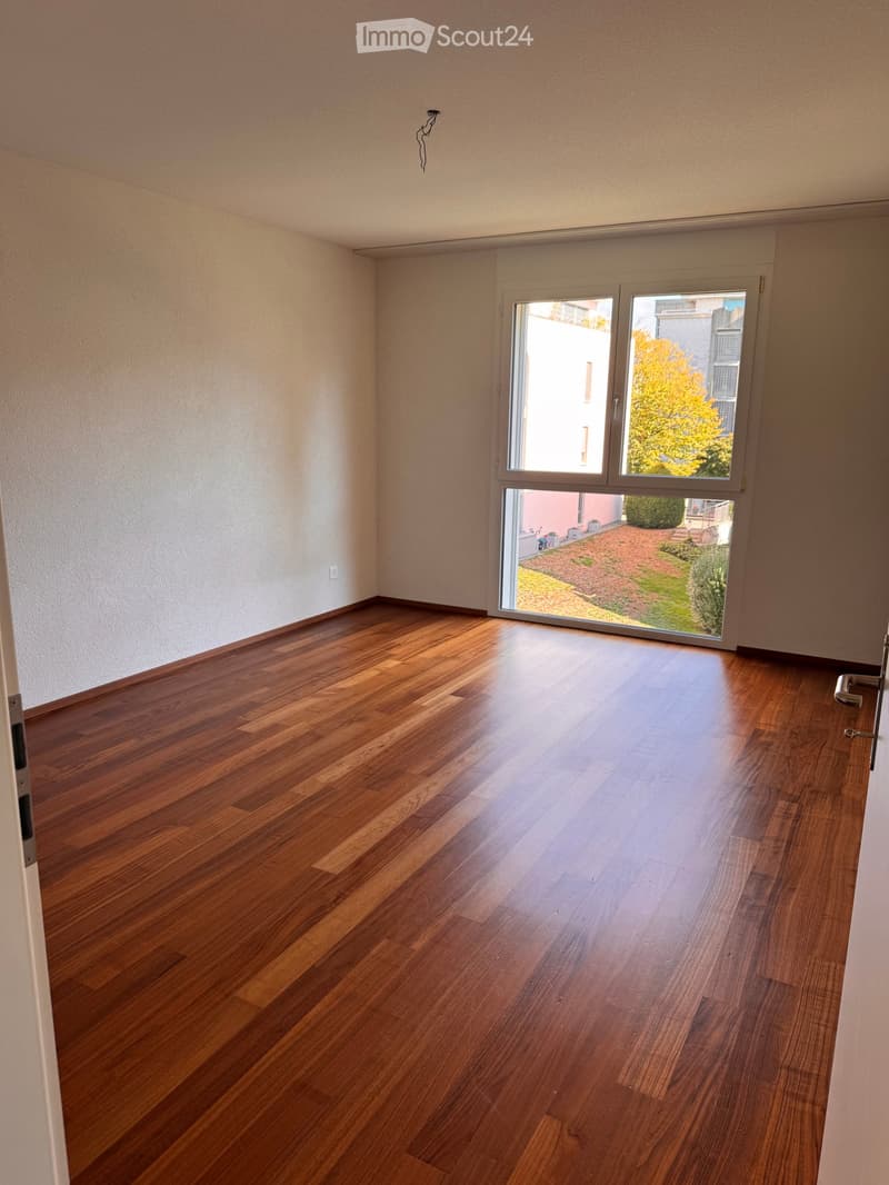Empty room with wooden flooring, large window with sliding glass doors leading to a garden area, light-colored walls, and a visible electrical outlet.