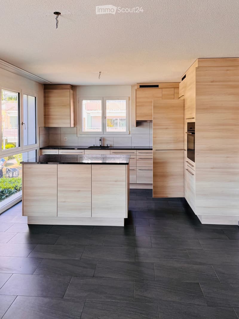 Modern kitchen with light wood cabinets, dark countertops, and a central island. Features include a sink, oven, and large windows offering a view of the garden.
