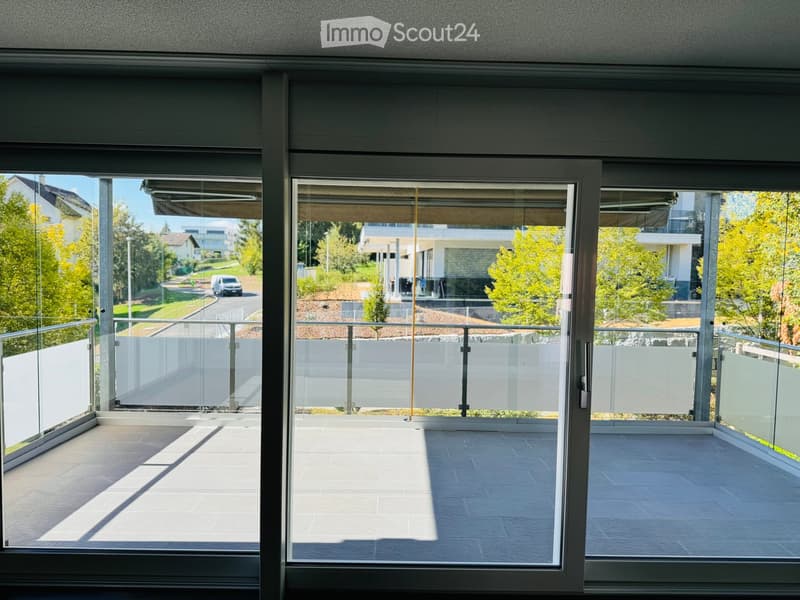 Modern balcony with large glass sliding doors, tiled floor, and metal railings, offering a view of a suburban neighborhood with trees and houses.