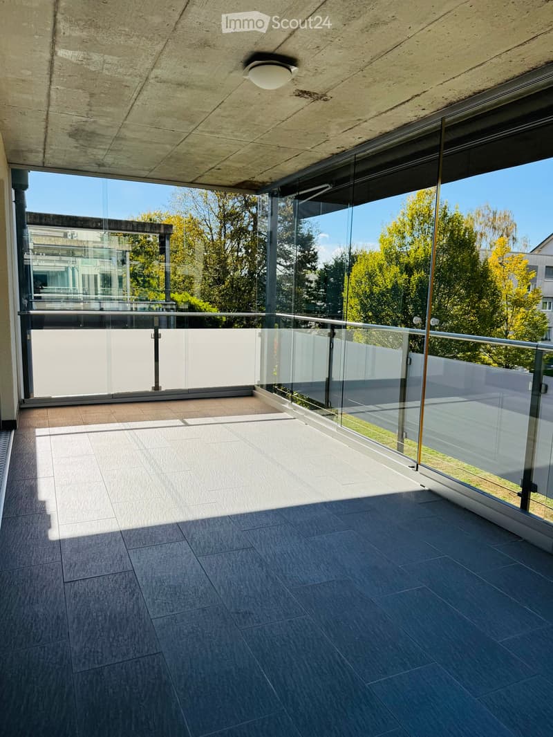 Modern balcony with glass railing, tiled flooring, and a concrete ceiling with a light fixture.