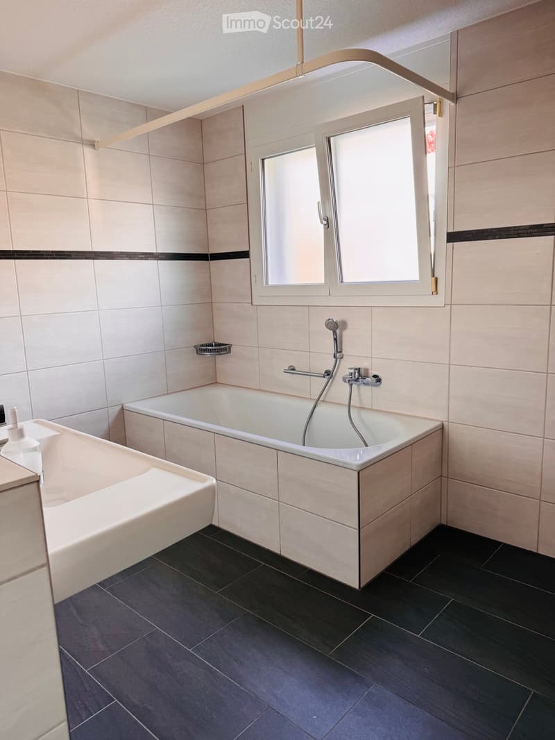 Modern bathroom with a bathtub, showerhead, and a large mirror above the sink. The walls are tiled in light beige with a black accent border, and the floor is dark grey tiles.