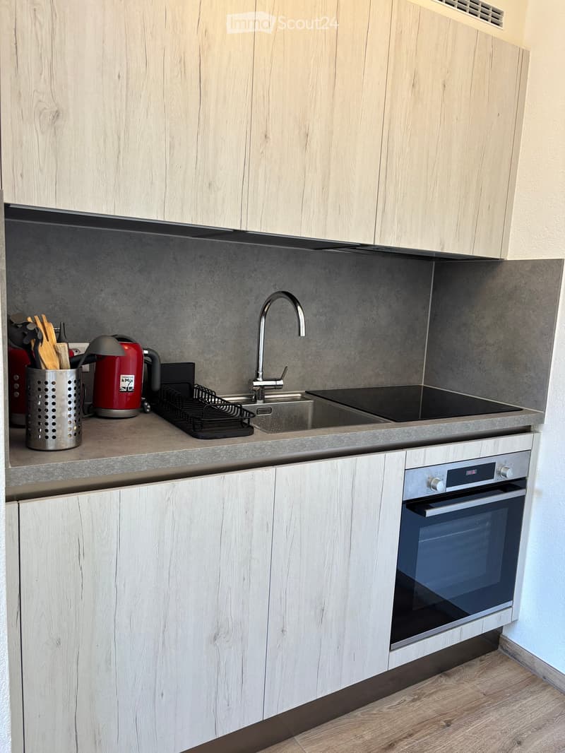 Modern kitchen with light wood cabinets, stainless steel appliances, and a grey countertop. Features include a sink with a modern faucet, an oven, and various kitchen utensils on the counter.