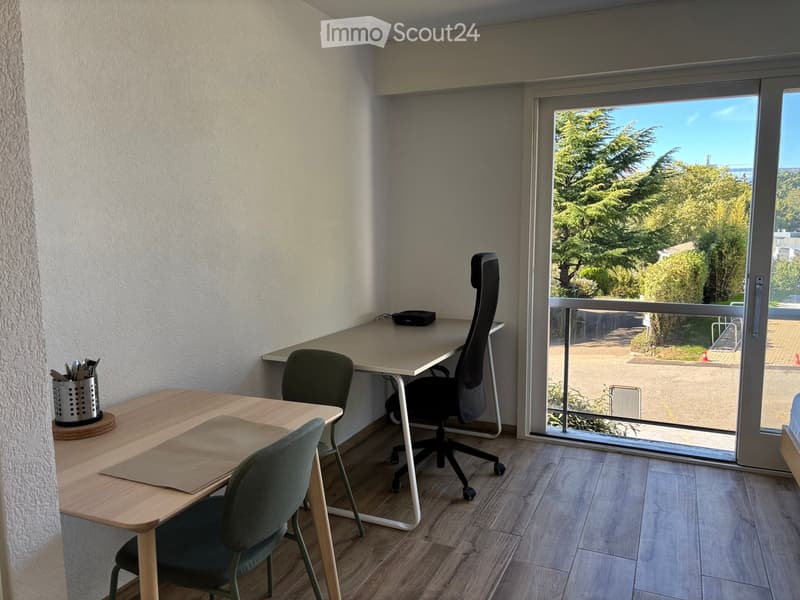 A modern, well-lit room with a desk, office chair, small table, and a laptop, featuring large windows with a view of greenery.