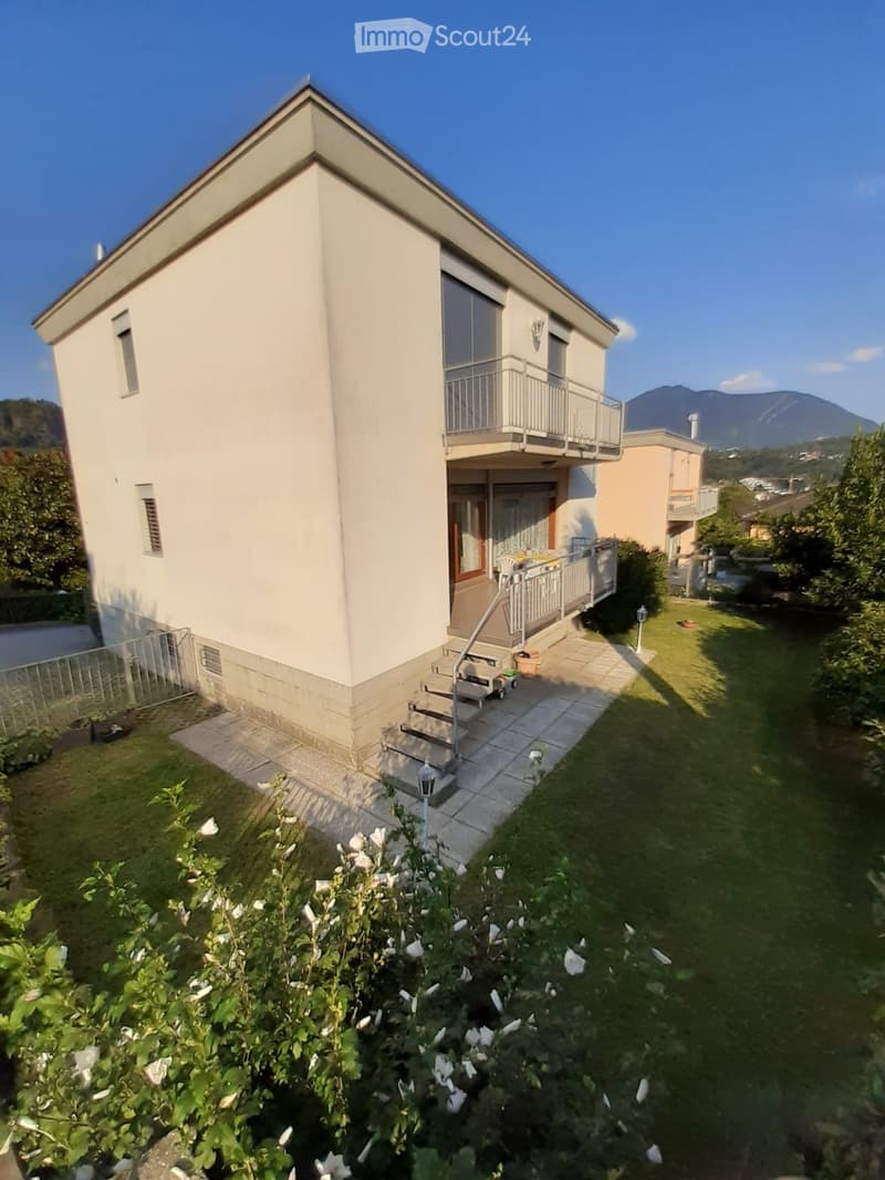 Two-story house with a balcony, steps leading to the entrance, and a well-maintained garden with flowers and shrubs.