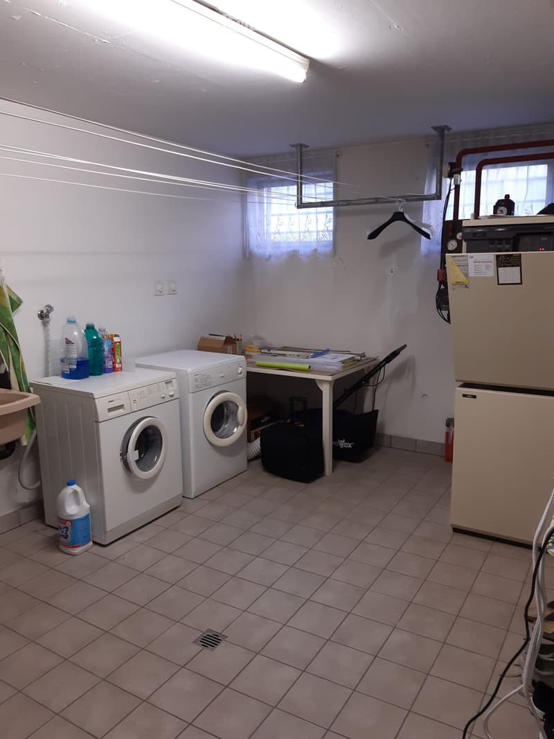 A small utility room with a washing machine, dryer, table, and refrigerator. The room has tiled flooring and white walls.