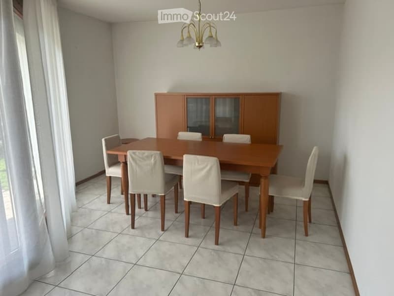 Dining room with a wooden dining table, six white chairs, and a wooden cabinet with glass doors. The room features tiled flooring and a chandelier hanging from the ceiling.