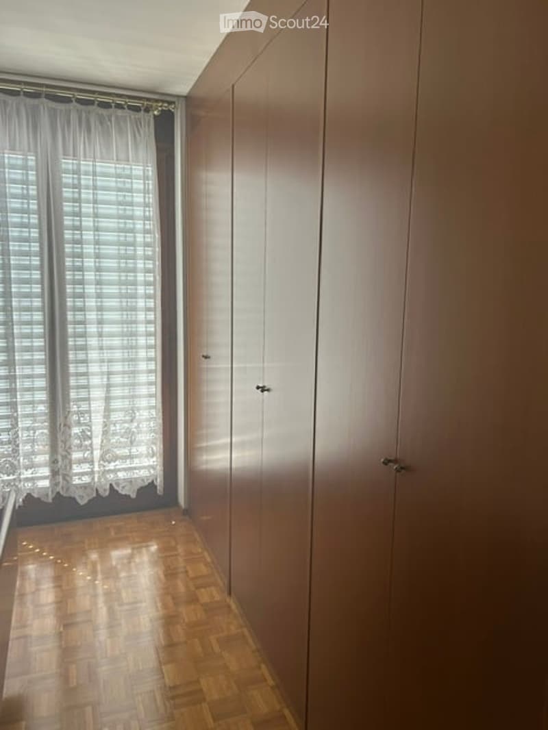 Empty room with wooden flooring, large built-in wardrobe, and window with white curtains.