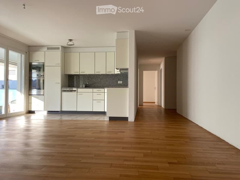 Empty room with wooden flooring, white walls, and a small kitchen area with cabinets and appliances visible in the background.