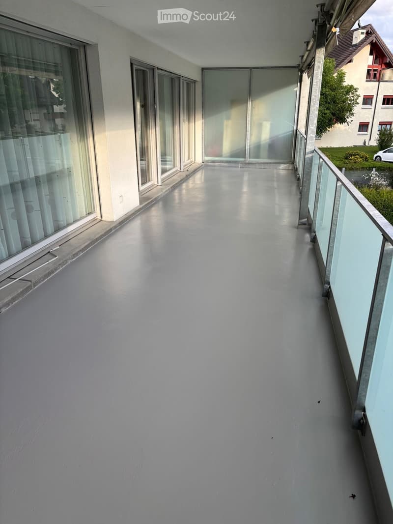 Modern balcony with smooth concrete floor, large glass sliding doors, and a glass railing, overlooking a residential area.
