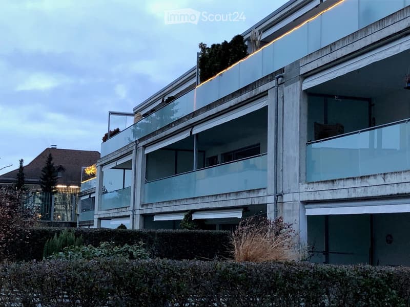 Modern multi-story building with large glass windows and balconies, surrounded by greenery.