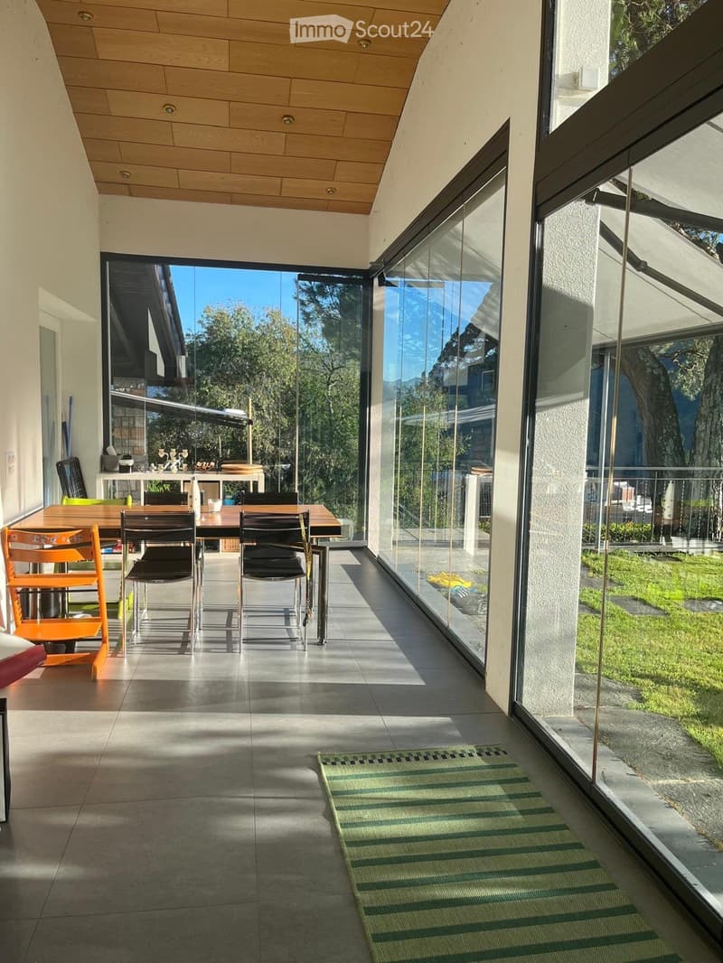 Bright, modern dining area with large glass walls, wooden table, and chairs, leading to a garden view.