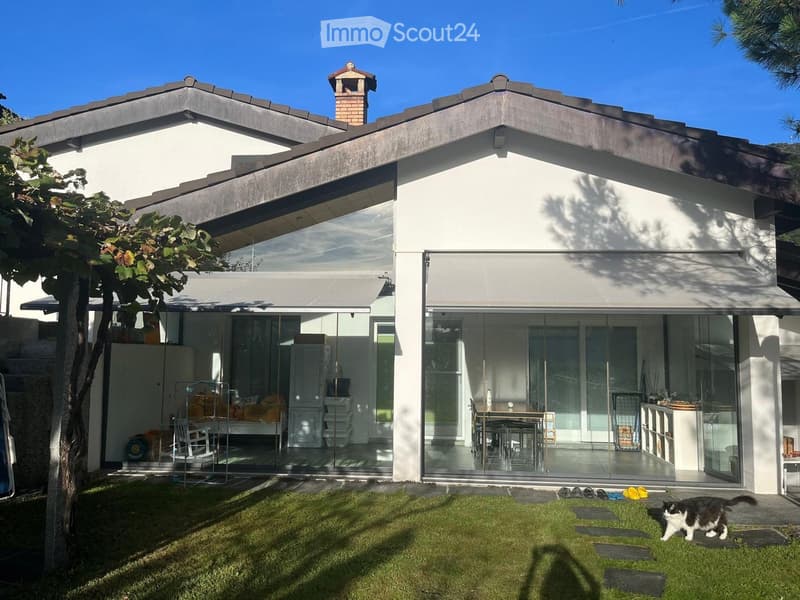 Modern two-story house with white exterior, large glass sliding doors, and a well-maintained lawn. The house features a raised ground floor and a small chimney on the roof.