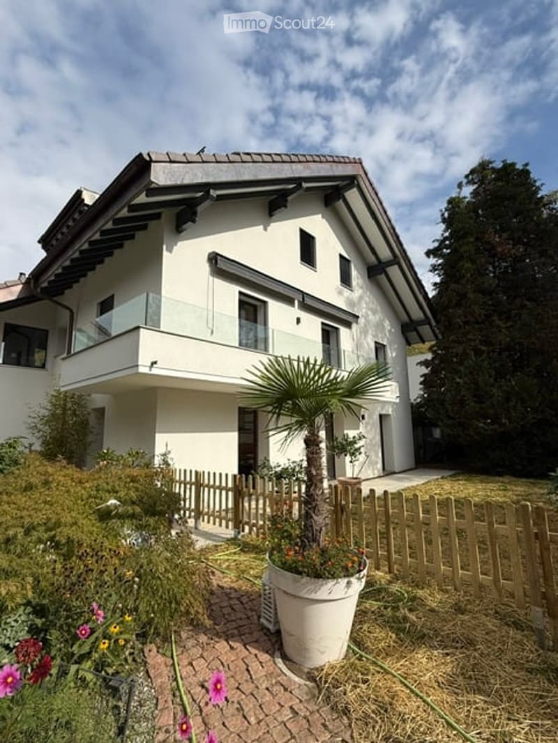 Modern two-story house with white exterior, large windows, and a balcony on the upper floor. The front yard features a wooden fence, potted plants, and a palm tree.