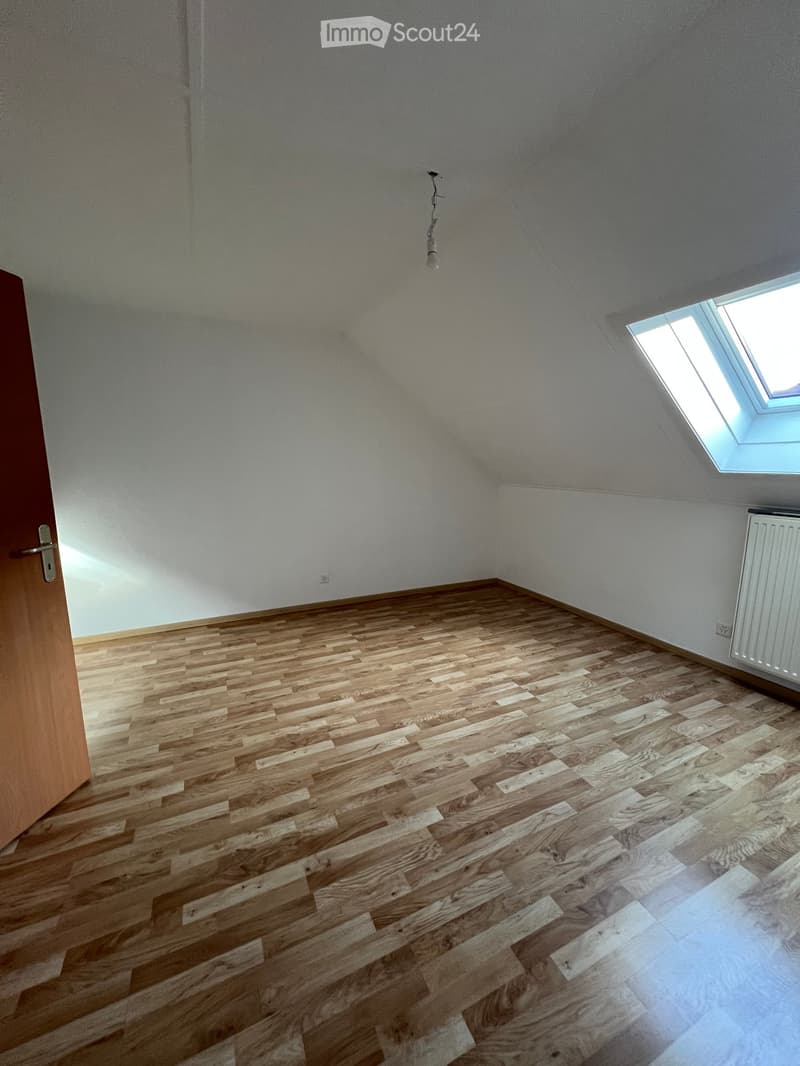 Empty room with wooden flooring, white walls, attic window, and a wooden door.