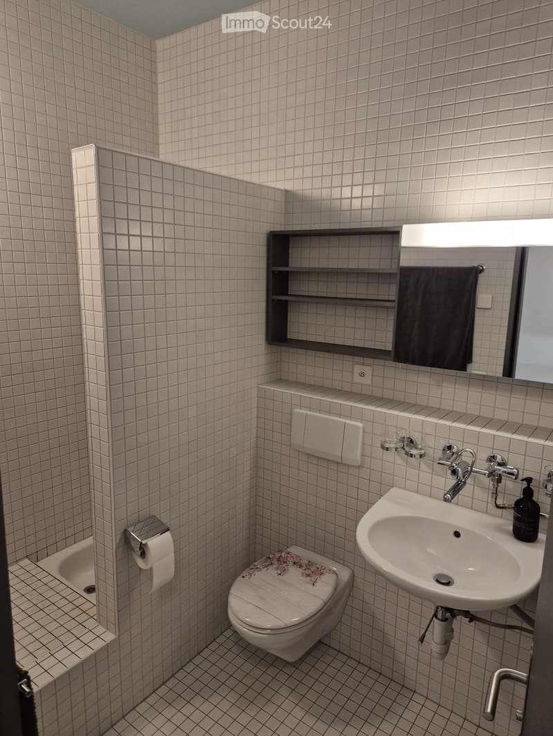 Modern bathroom with white tiled walls and floor, featuring a toilet, sink, mirror, and built-in shelf with a towel. The sink has a modern faucet and a soap dispenser, and a small shelf holds a towel and other items.