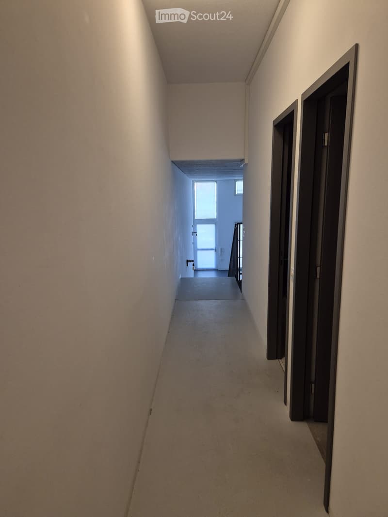 Empty hallway with beige walls, carpeted floor, and two closed doors on the right side. A window at the end provides natural light.