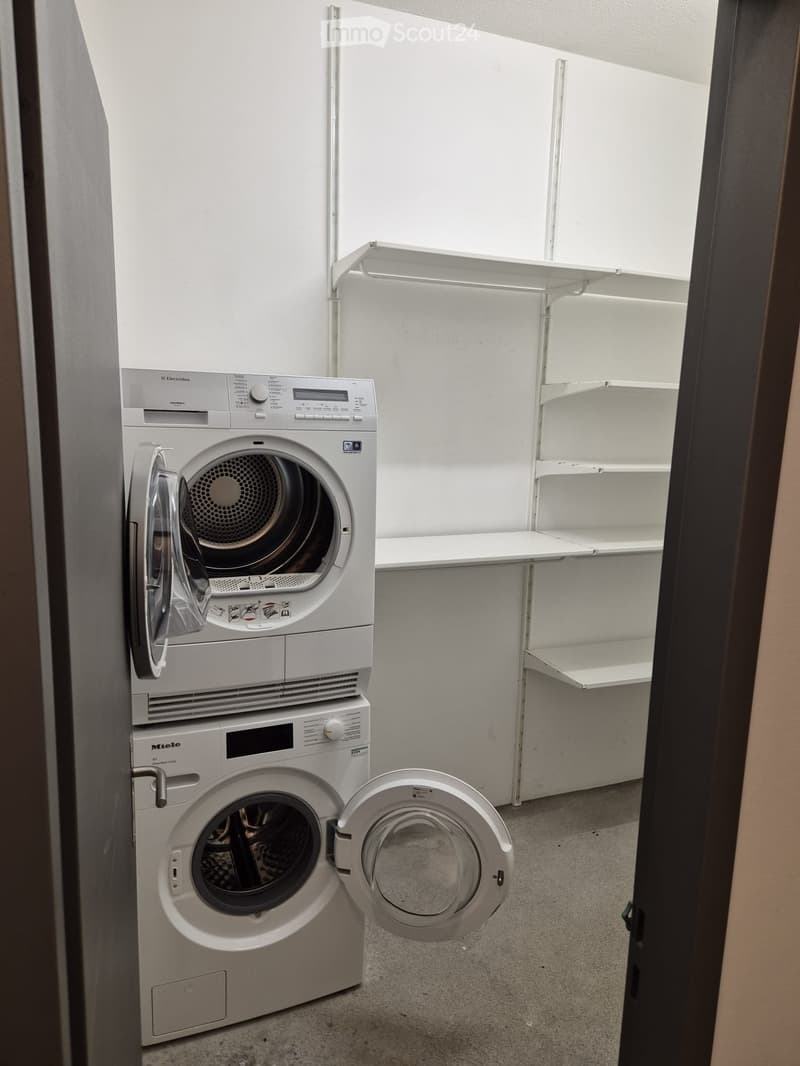 A small, well-lit room with a washing machine and dryer stacked vertically, and a built-in shelf unit on the wall.