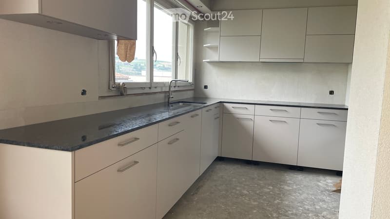 Modern kitchen with white cabinets, black countertop, and a sink with a faucet. The floor is unfinished concrete, and there is a large window with a view.