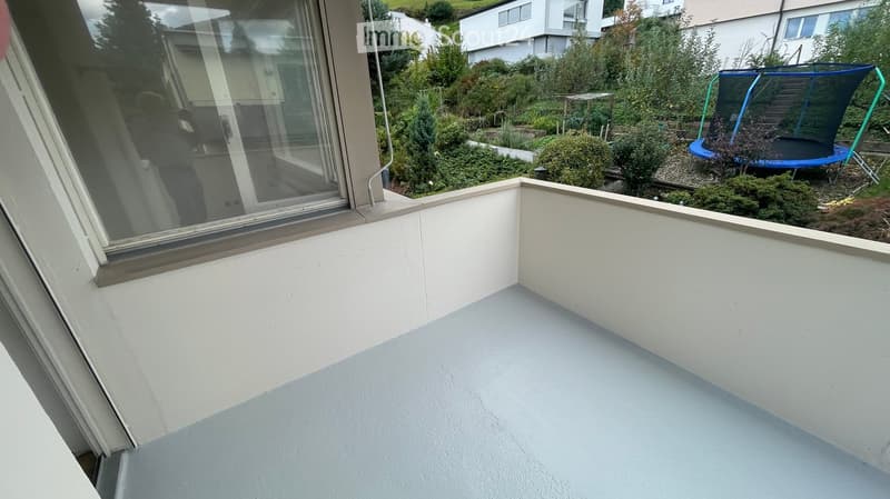A modern, white-painted balcony with a large window overlooking a garden area with bushes, trees, and a trampoline.