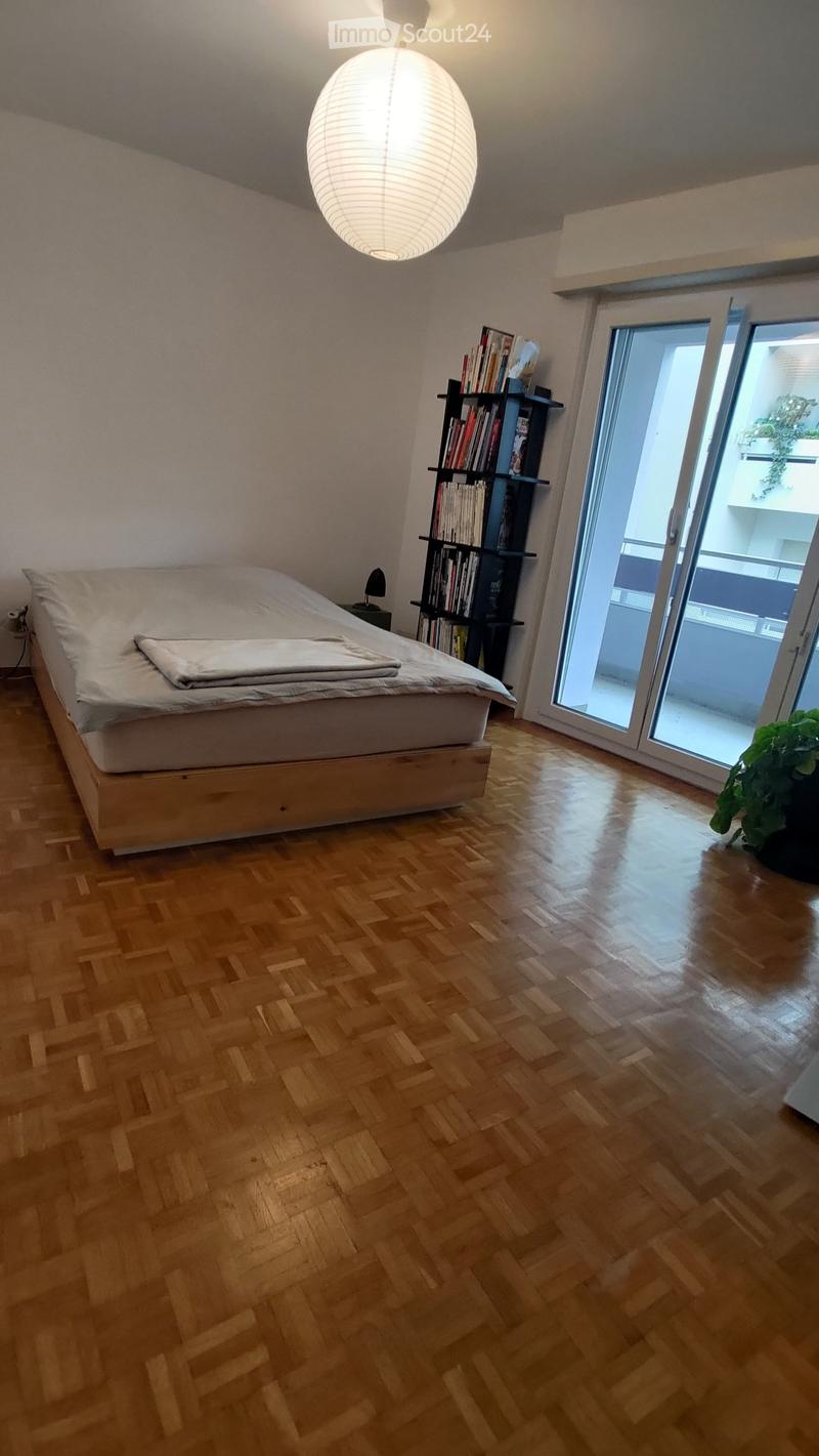 Modern bedroom with wooden herringbone flooring, a large bed with white bedding, a bookshelf, a table lamp, and a potted plant near the sliding glass door leading to a balcony.