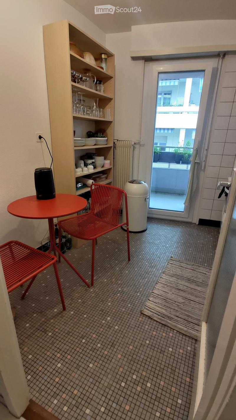 A small, furnished room with a red table, red chairs, a shelf with dishes and cups, and a view of a balcony through a glass door.
