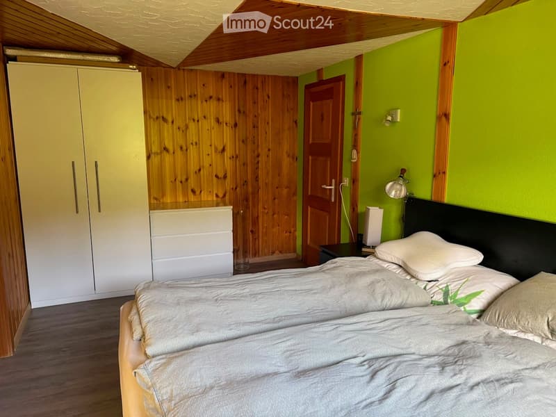 Bright bedroom with green walls, wooden floor, bed with white bedding, nightstand with lamp, wardrobe, and drawer unit.