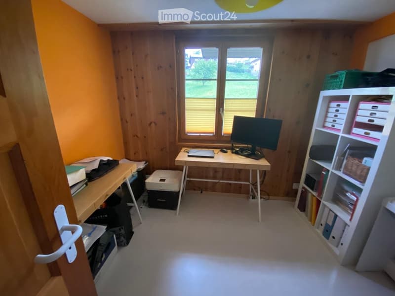 A small, well-lit room with wooden walls and a large window. Contains a desk with a laptop, monitor, and printer, along with a shelf holding various items.