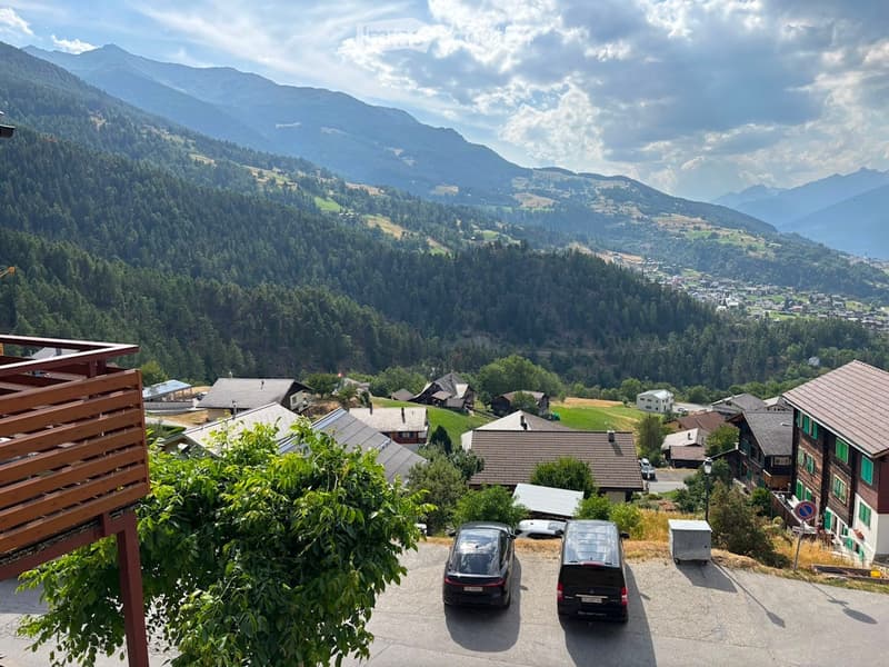 Panoramic view of a valley with houses, trees, and mountains in the background.