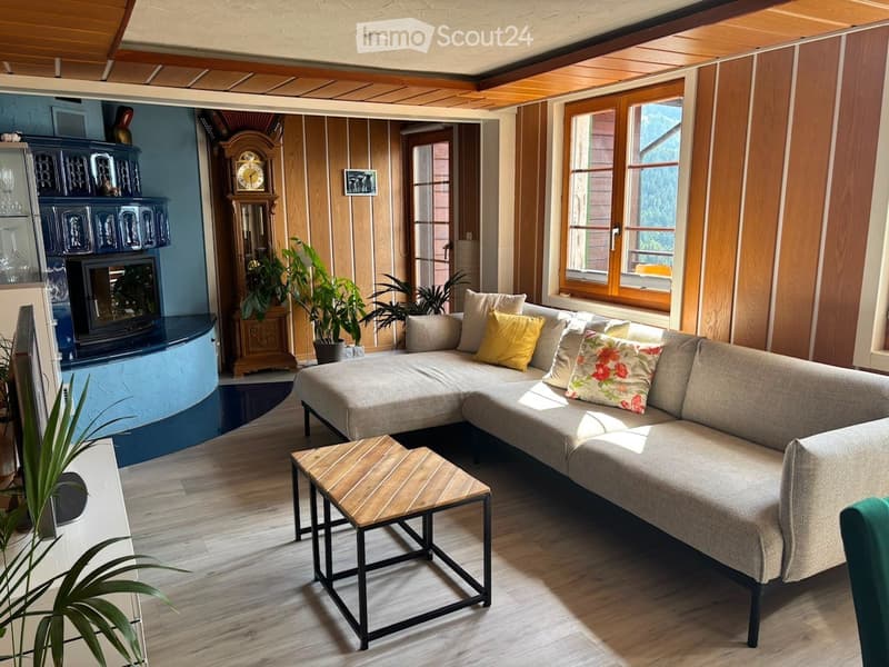 Cozy living room with a grey sectional sofa, wooden coffee table, fireplace, and potted plants. Large windows provide natural light.