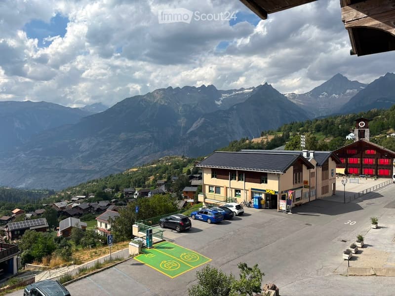 View of a parking lot with several cars, surrounded by buildings and lush greenery, with a backdrop of snow-capped mountains.